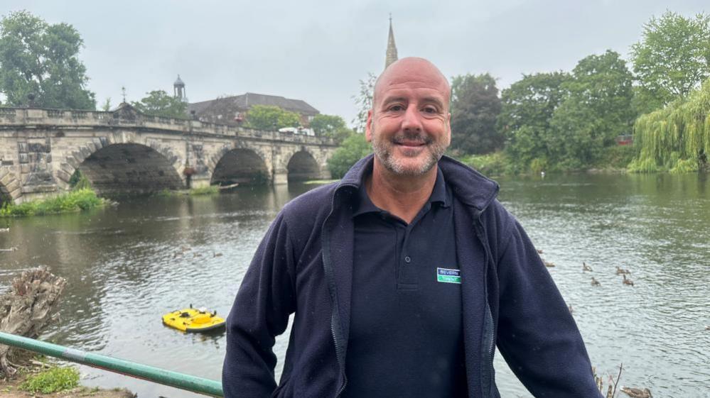A bald man standing in front of the River Severn. A large stone bridge with four archways his behind him in the distance as well as a church spire. The man is wearing a navy blue polo shirt and a navy fleece. He is smiling and leaning on a green railing. There is a bright yellow paddle board on the river behind him to the left. 