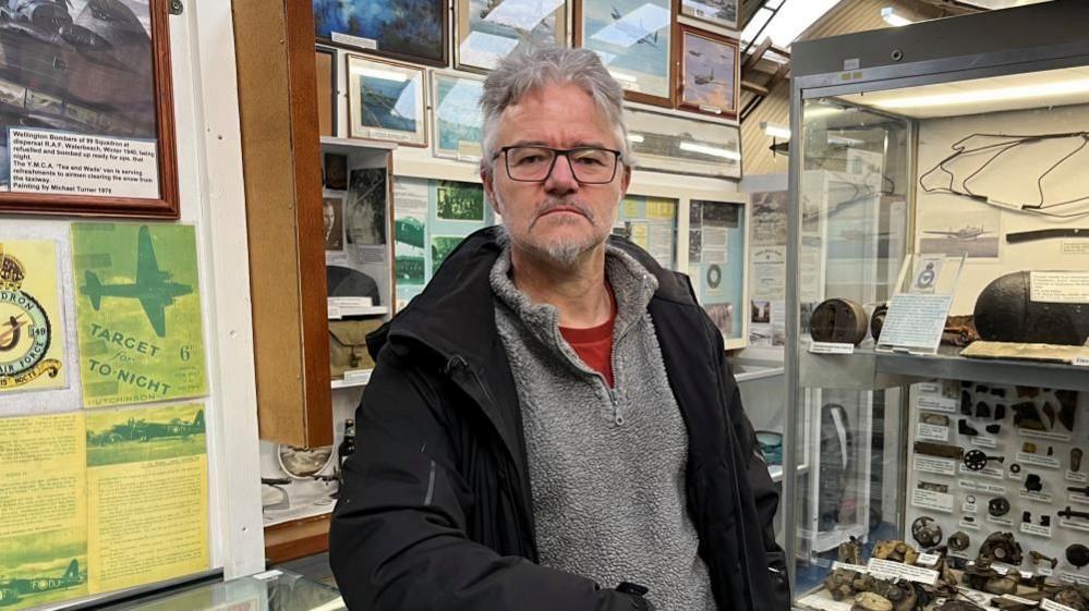 Rob Spray, wearing a grey fleece and black jacket, is looking at the camera with a range of framed photos of World War Two planes behind him. He's standing in the Norfolk and Suffolk Aviation Museum near Bungay.