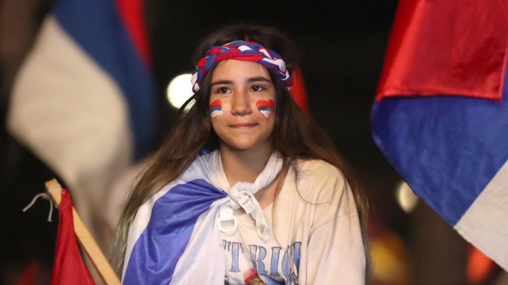 A girl with a heart in the colours of the Broad Front flag painted on one cheek and the Broad Front flag on the other smiles after the victory of Orsi was announced. She has a Broad Front flag tied to her neck and is waving another one in her hand. 