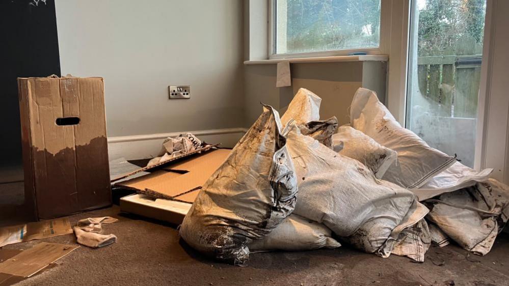 A pile of sandbags inside a room that has been flooded - there is dirt everywhere and there are soggy cardboard boxes. 