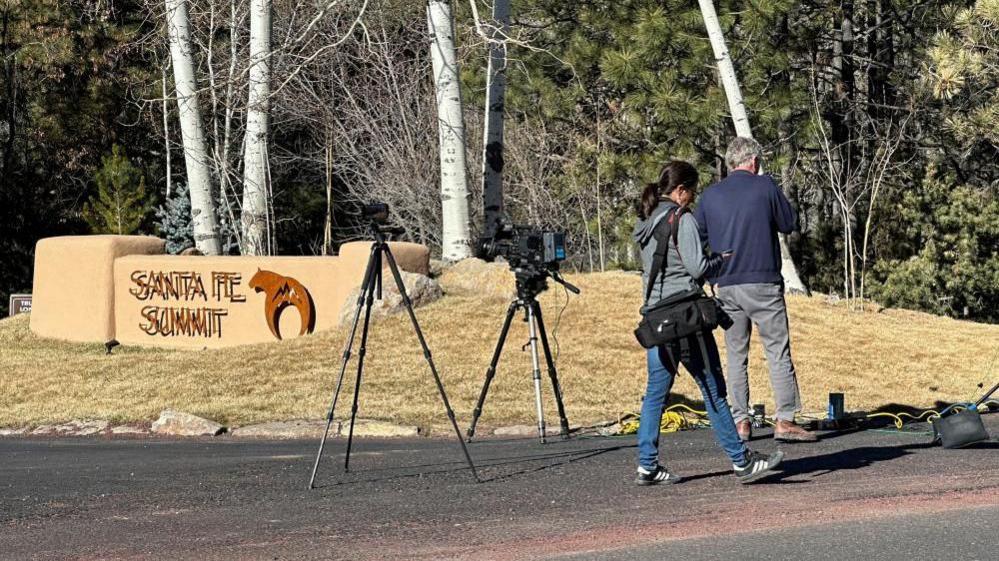 Reporters gathered outside the gated community where Hackman and his wife died. Two reporters wear denim jean pants, and stand near tripods holding up cameras. They face a sign that says 'Santa Fe Summit'