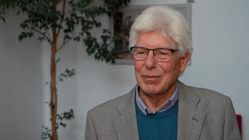 Councillor Paul Heinrich has white hair, wears glasses and is wearing a blue and white shirt, turquoise jersey and checked jacket. He is in a room painted white, with a potted tree in the background.