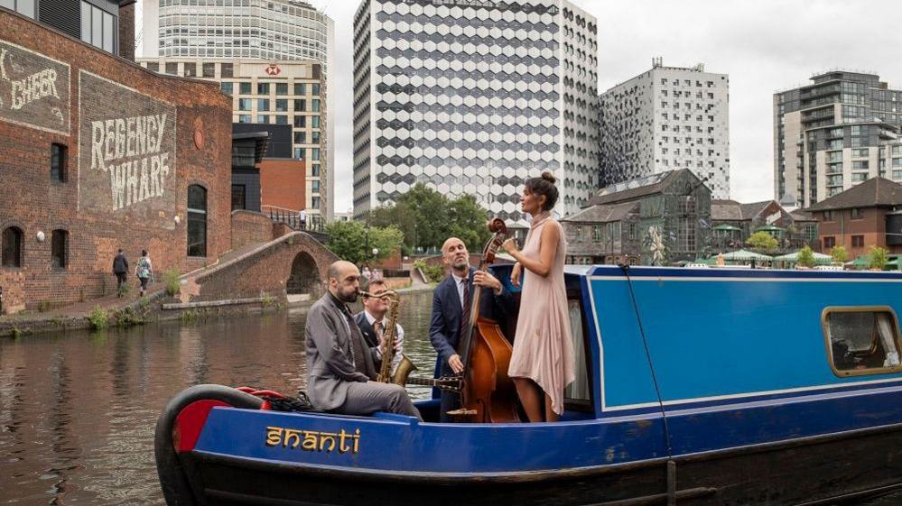 Three men and a women are standing on the edge of a blue canal boat. They are performing together using jazz instruments like the trumpet and double bass. The boat is sitting on the canal with high-rise buildings in the background