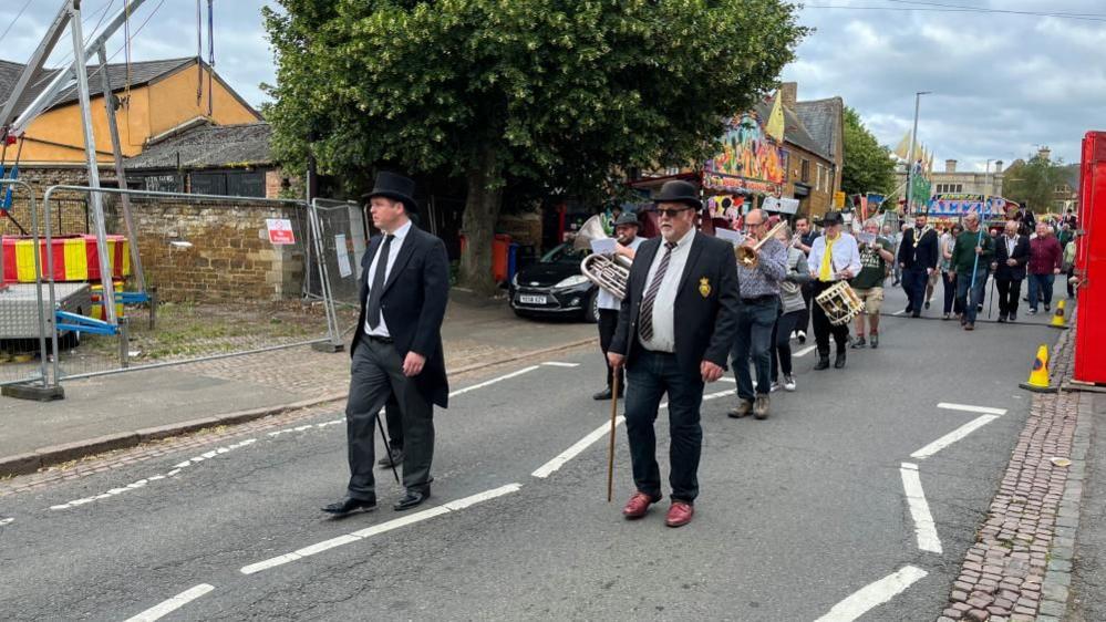 Two smartly dressed men lead a procession with a band and several people behind them on a road.