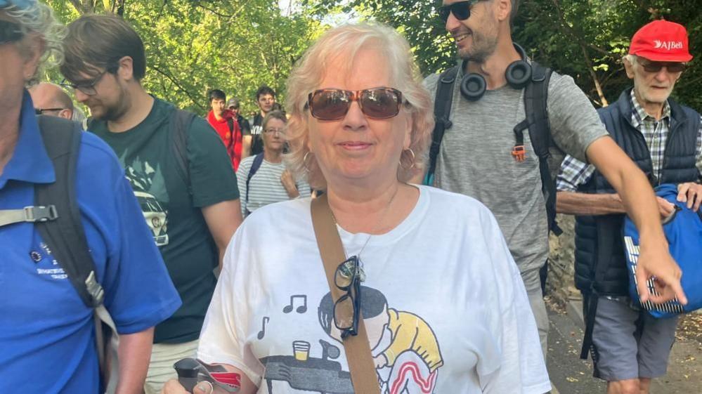 A woman wearing a music-themed T-shirt and carrying a walking pole walks among a large group of people on a footpath, with trees in the background. She is wearing sunglasses with spectacles hanging from a necklace.
