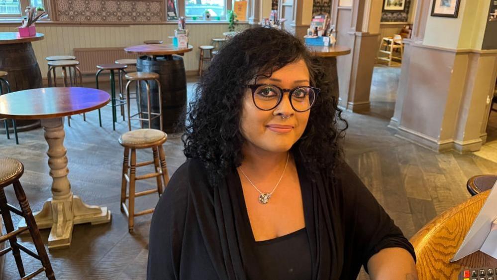 Shumi Plastow with long dark hair and big glasses, wearing a black dress and a silver necklace and sitting in a pub.