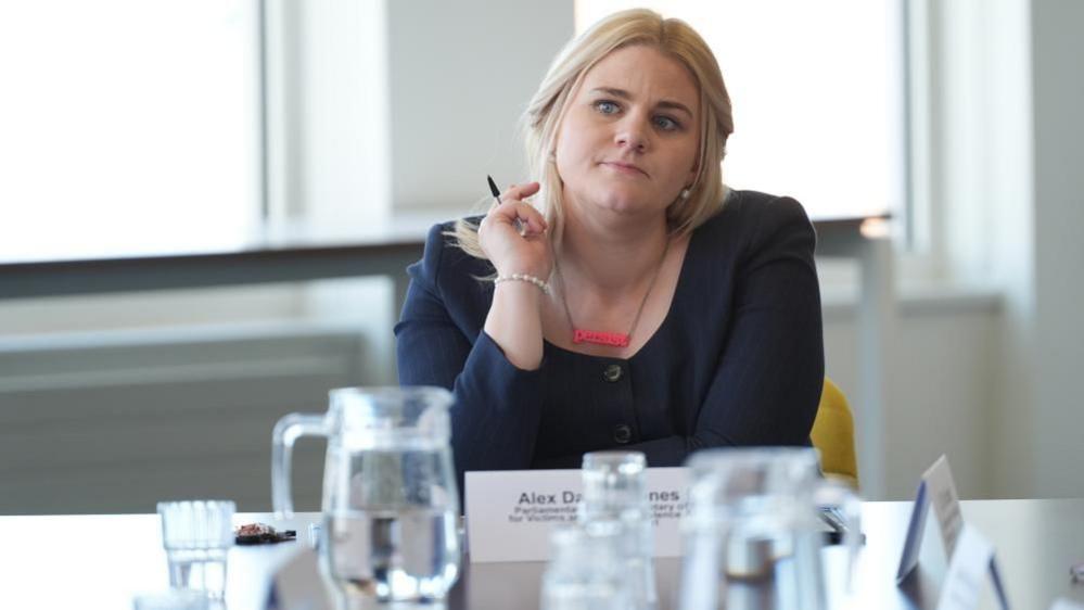 Alex Davies-Jones is sat on a chair and is holding a pen in her right hand. The table in front of her has a jug of water and several glasses. She is wearing a dark coloured top.
