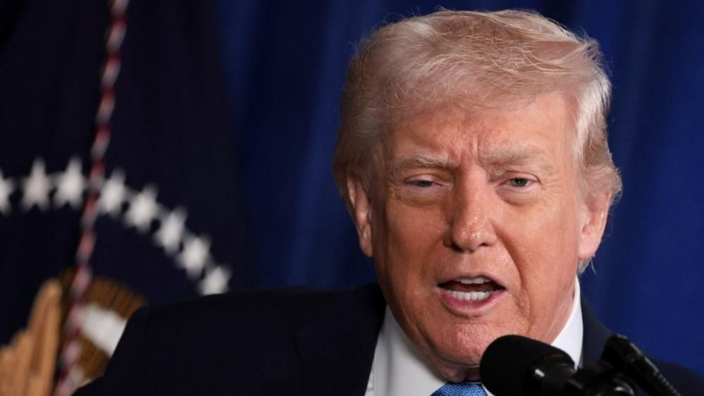 Donald Trump stands in front of a blue background and speaks at a microphone. He is wearing a black suit, white shirt and blue tie. 