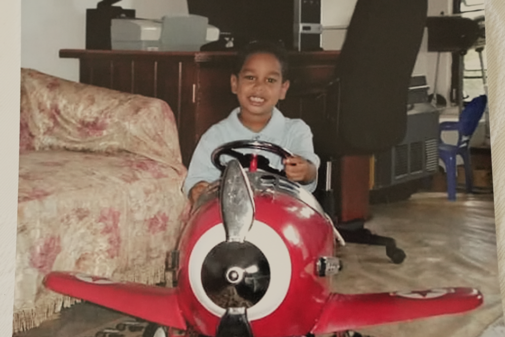 Shawn Seesahai as a young boy, sitting in a big red aeroplane toy while holding its steering wheel; he is sat in front of a desk with a computer and computer chair, and a sofa is to his left