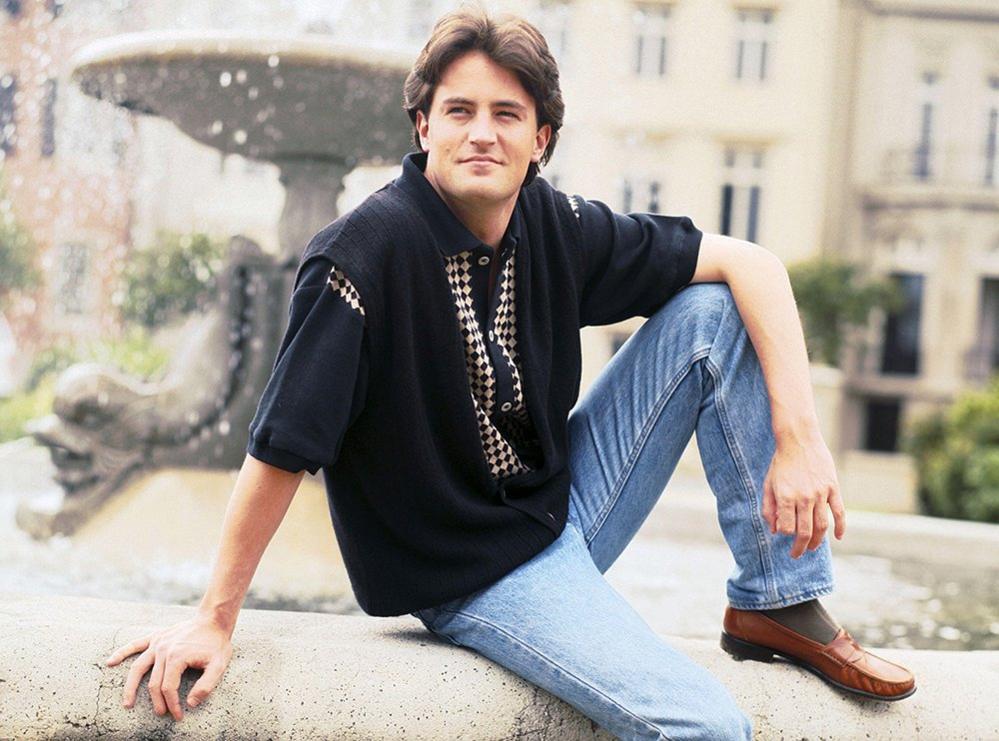 Matthew Perry poses for a portrait on a fountain, wearing blue jeans and a polo shirt