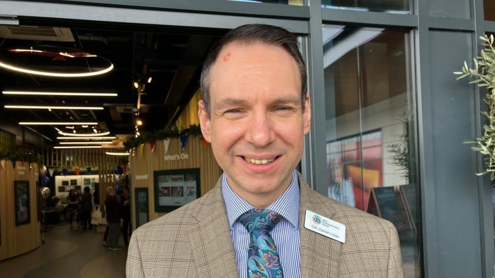 Dan Lister with short dark hair wearing a brown jacket and blue and white striped shirt with blue tie. He is standing in the foyer of a large building at Rushden Lakes.