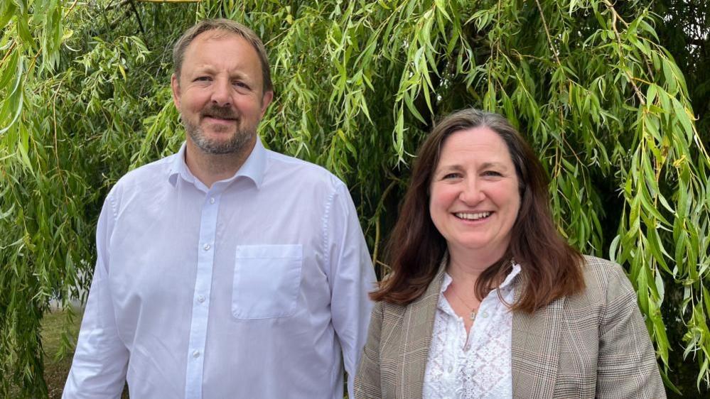 Toby and Julia stood side by side in front of a willow tree, both are smiling. Toby is wearing a white button-up shirt and Julia is wearing a beige tweed blazer with a white blouse.