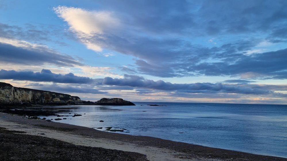 Marsden Bay looking out to the North Sea at dusk. The sky is still light blue in parts but the clouds are turning grey. The sea looks calm and cliffs can be seen to the left of the coastline.