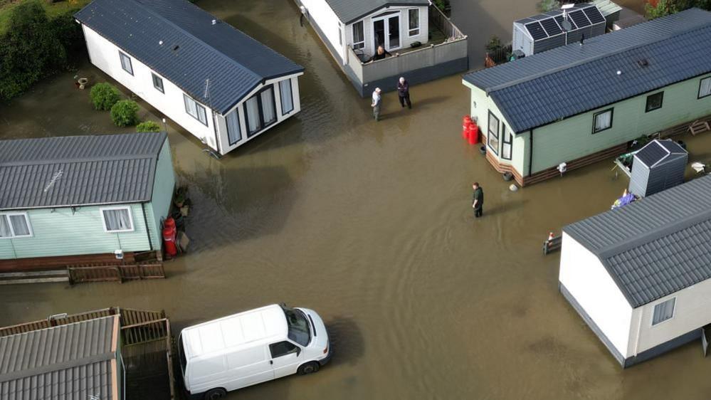 Mobile homes green and white in colour, with grey roofs, surrounded by water, pictured from the air. A few people are visible standing in the water and there is a white van parked to the bottom left of the picture.