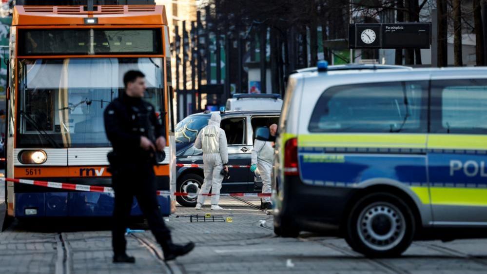 In focus, two police in white hazmat suits walk by a black car. In the foreground, obscuring the car and one policeman, is a police van and a uniformed officer who walks over the tramline