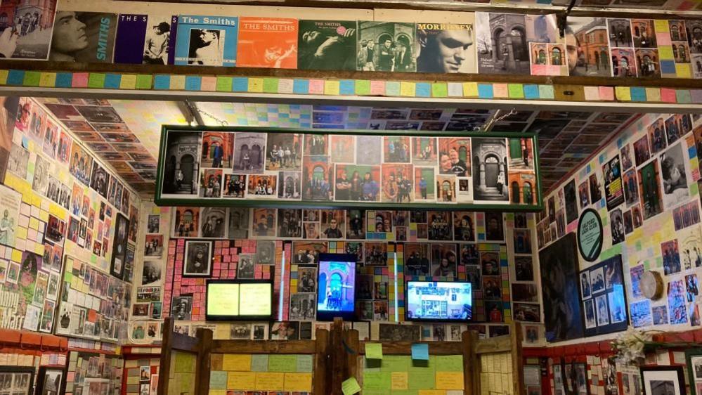 A photo of the inside of the Salford Lads Club shows walls covered in postcards, posters, post-it notes and photographs. 