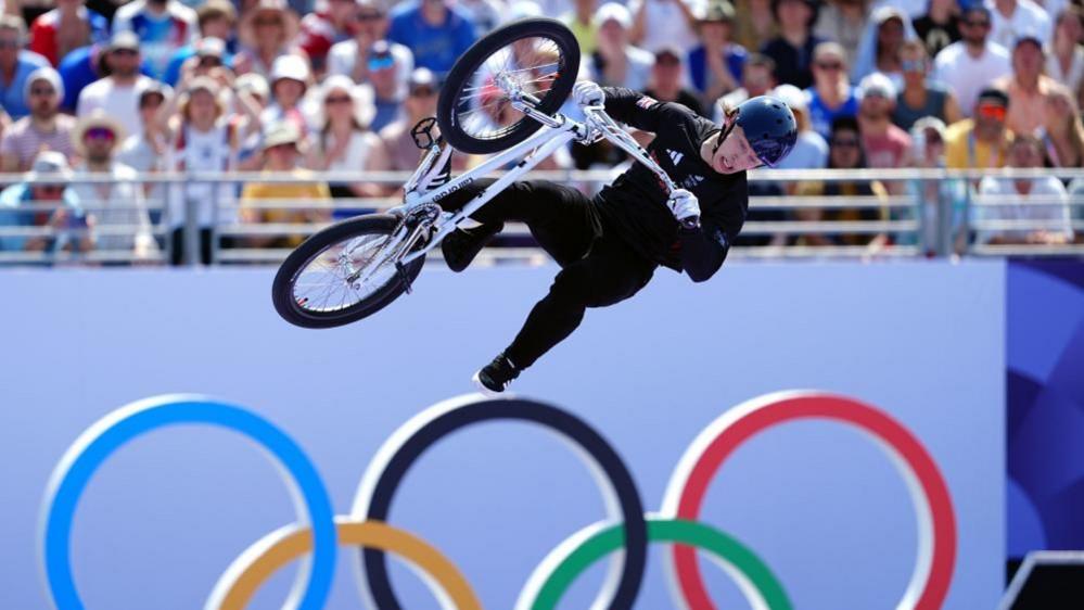 Kieran Reilly dressed in black with helmet and on his bike, which is airborne above an Olympic symbol