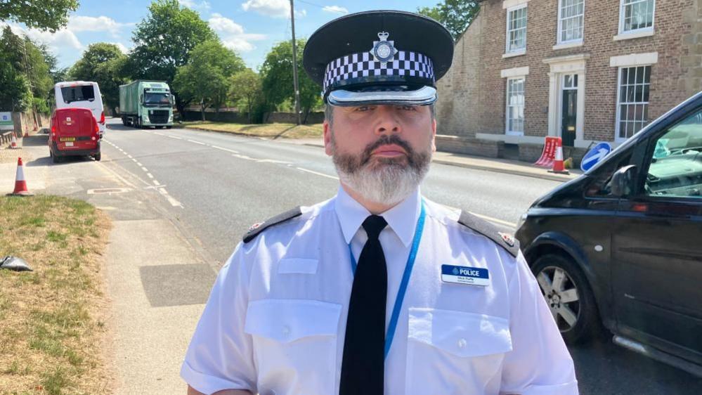 A police officer wearing a white shirt and black tie and black police cap looks into the camera with a neutral expression on his face. He is standing next to a road, with traffic passing in the background.