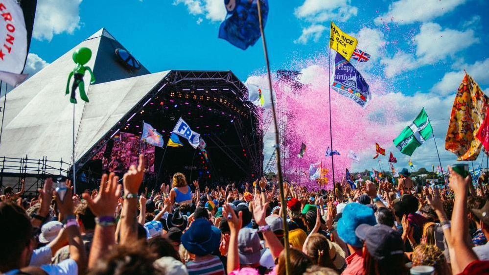 People with raised hands enjoying the performance on the Pyramid stage with pink confetti in the air and people holding flags with happy birthday and other messaging