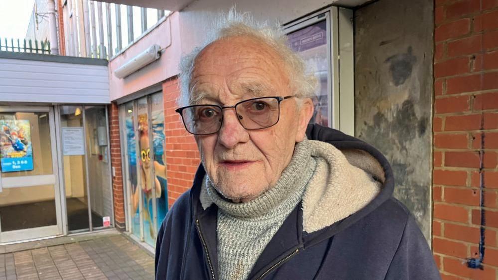 A man with glasses, and roll-neck grey jumper. He is also wearing a hoodie. He is stood in front of a leisure centre.