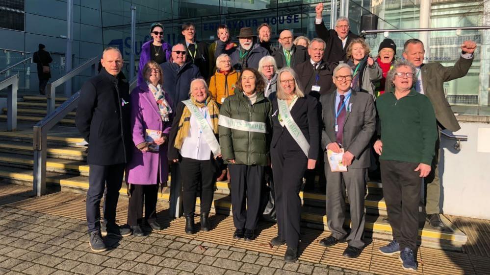 A group of people protesting outside Suffolk County Council. They are a mix of local councillors and activists. There are a mix of men and women. They are wearing coats and smart clothing. 