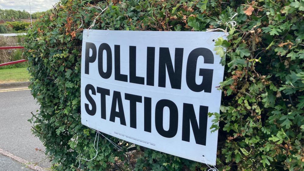 Stock image of a sign reading 'polling station' with black capitalised lettering on a white background, against a hedge