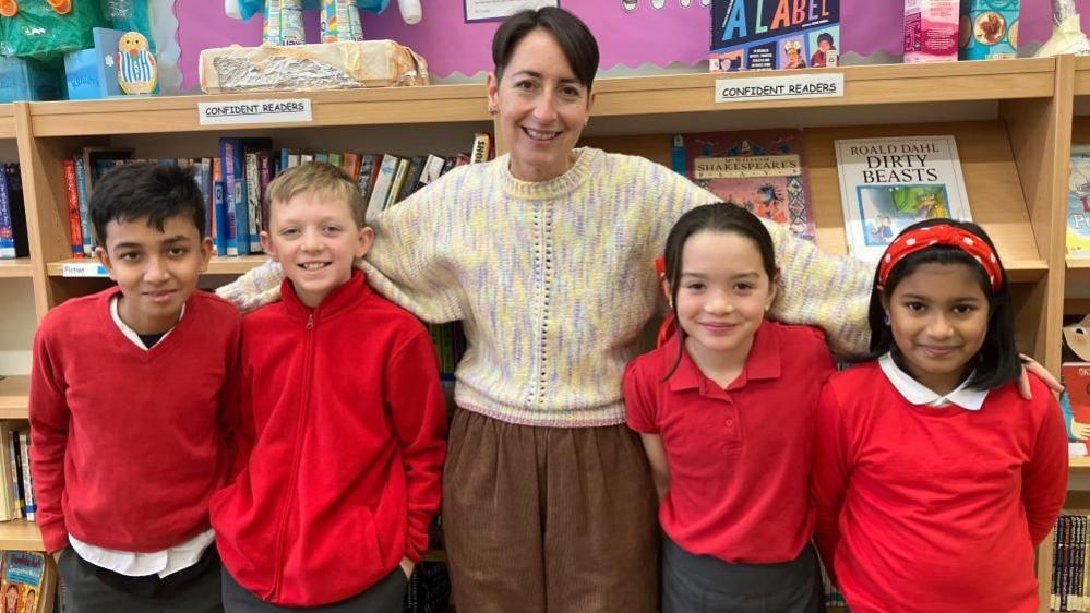 Deputy headteacher of Fishergate Primary School Dani Rees stands in the library with a mixed gender group of Fishergate Primary School pupils. Ms Rees wears a knitted jumper with purple, yellow and pink threads, and brown cord trousers. The pupils wear red jumpers or polo shirts.