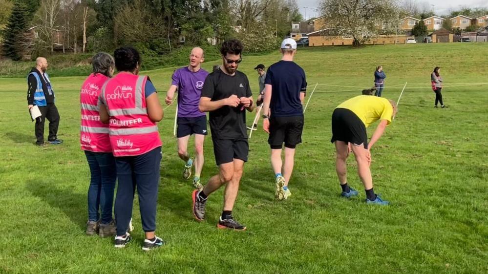 A field with trees and houses in the background. There are ropes guiding runners to a finish line, where two women in pink hi-vis vests are taking their position tokens. One man in a purple T-shirt and black shorts is handing his token over.  A man with black hair and beard is checking his position ticket, and a man with a yellow T-shirt and black shirts is bent double after finishing.