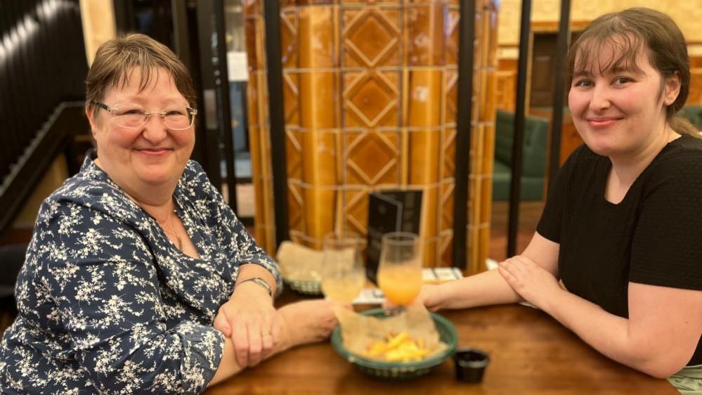 An older and a younger woman sit across a table from each other. They are each holding a glass of orange liquid and a plate of chips is on the table between them. 