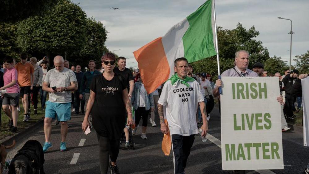 Protesters march holding banners and the Irish flag during the demonstration on July 19, 2024 in Dublin, Ireland. One man is holding a banner saying "Irish Lives Matter" as well as an Irish flag. The man next to him is wearing a t-shirt saying "Coolock says no" and wearing an Irish flag around his neck.