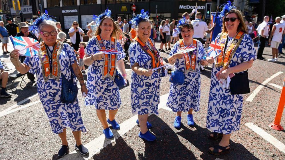Five ladies in matching blue and white dresses wearing orange sashes. 