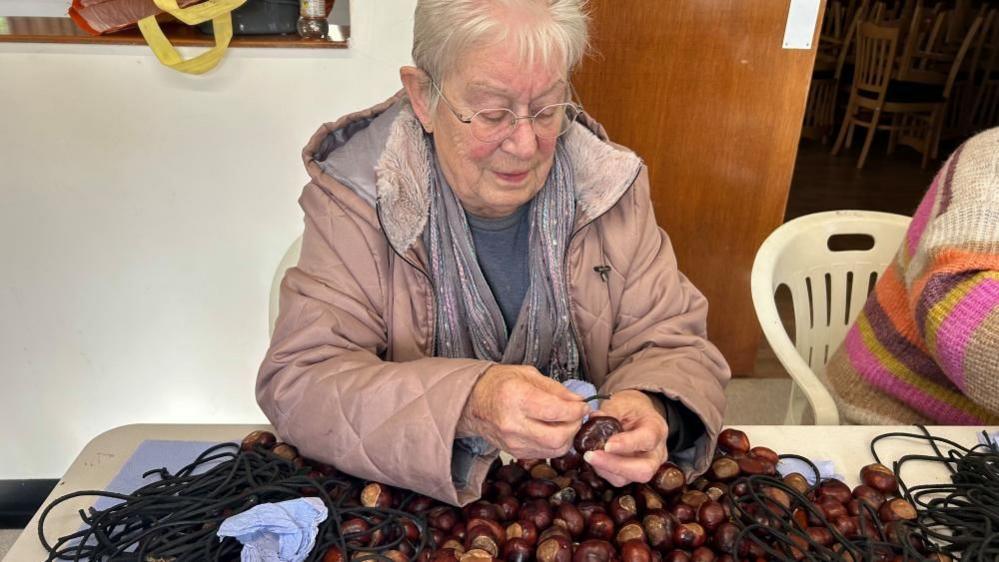 Sue Howes with short white hair wearing a pink anorak and peeling a conker. There is a pile of conkers in front of her on a table. She is in a room with a wooden door and sitting next to another woman in a colourful sweater who is selecting conkers.