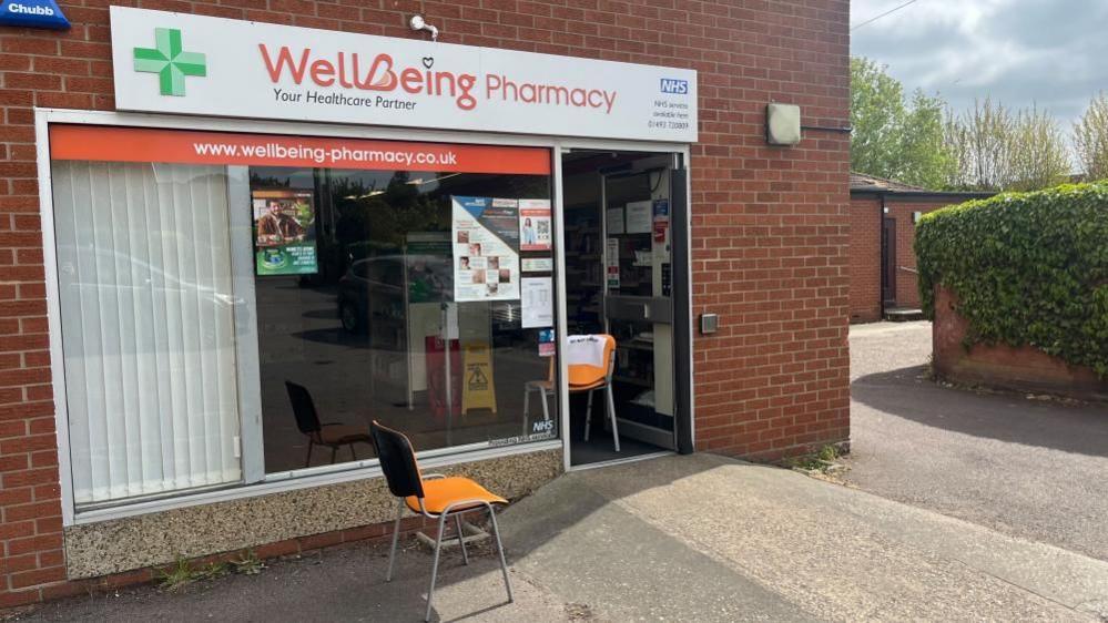 The Wellbeing Pharmacy, with it's pharmacy green cross logo on the left of the signage and NHS logo at the right of that sign, above the shop window and door. There is an orange chair in the doorway, and another outside near to the access ramp to the front door. There are blinds against part of the aluminium-framed window. The building is made of red brick, and part of the boundary wall is covered in ivy, with trees growing behind the buildings and a clouded sky appearing in the upper right of the image.