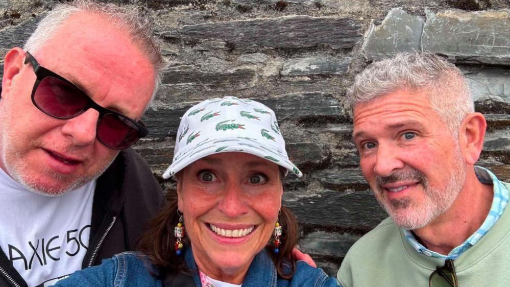 Mike, left, has short grey hair and is smiling at the camera. He is wearing red tint sunglasses. He is wearing a white t-shirt and a black zip-up hoodie. Christine, middle, is smiling at the camera and has a white Lacoste cap on. She has brown hair. Terry, right, is smiling at the camera and has grey hair and a grey beard. He is wearing a pale green sweatshirt with a blue checked shirt underneath.