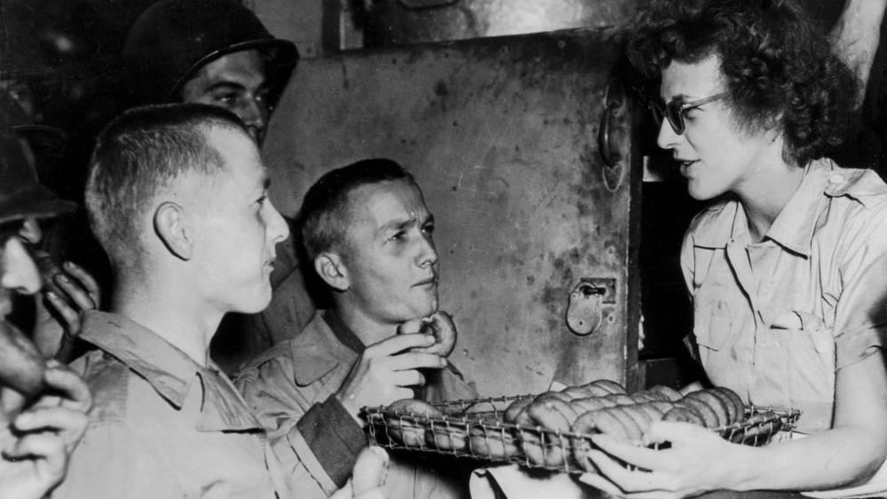 Two American soldiers, who are brothers, sample doughnuts offered by a member of an American Red Cross Clubmobile in Normandy (summer 1944). She is on the right with the tray in front of her. The two men are on the left and looking up at her, holding a doughnut each. 