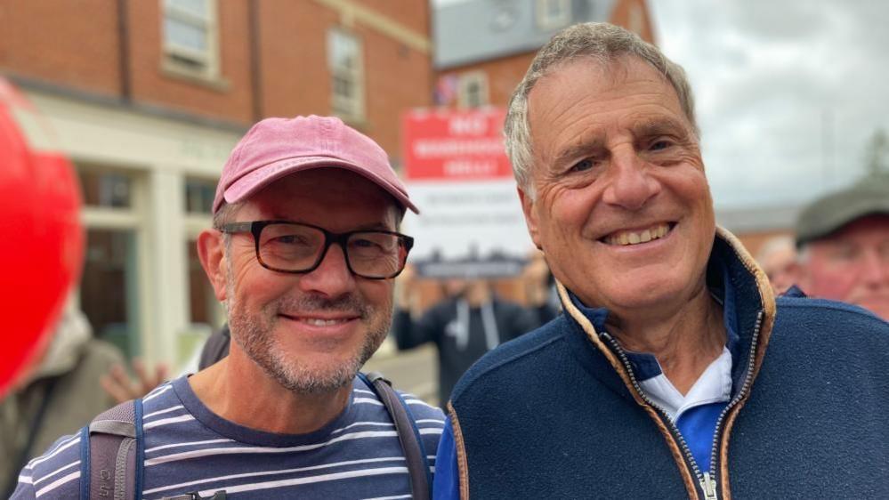 Graham with short grey beard and brown glasses wearing a pink cap and striped blue and white shirt. Geoff with short grey hair wearing a blue jacket with brown trim