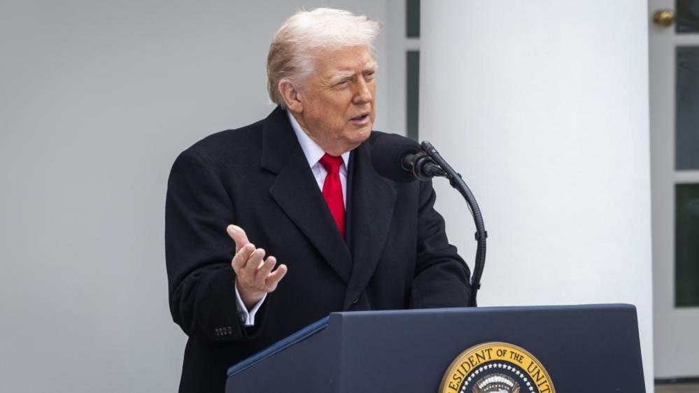 Donald Trump wears a black suit and red ties as he speaks at a podium with the presidential seal on the front