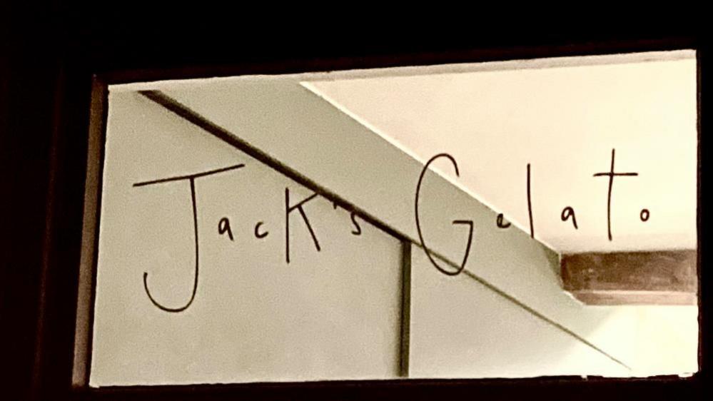 Sign on a fanlight at a Jack's Gelato ice cream shop in Cambridge. The words "Jack's Gelato" are written in black handwriting on a window. Part of a wall and ceiling, and a beam, are visible through the glass. 