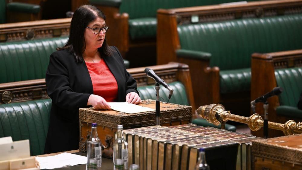 Rachel Hopkins wearing a black jacket over a red dress, asking a question in the House of Commons, she has dark long hair, glasses and has a necklace on. The seats are green leather, she is talking into a microphone and is holding a sheet of paper. 