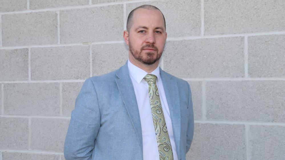 Det Ch Insp Nick Gardner: a man with short dark hair and dark beard with his hands clasped behind his back. He is wearing a pale blue jacket, pale shirt and yellow patterned tie.