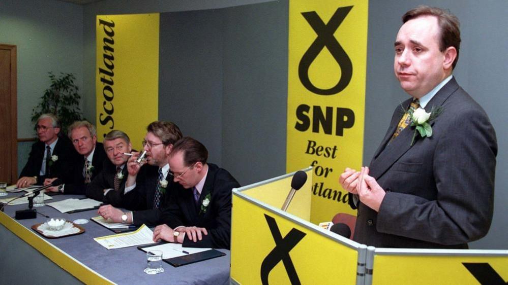 Alex Salmond stands at a podium with SNP branding in 1997. There are yellow SNP banners on the walls and five other senior party figures are sitting at a table.