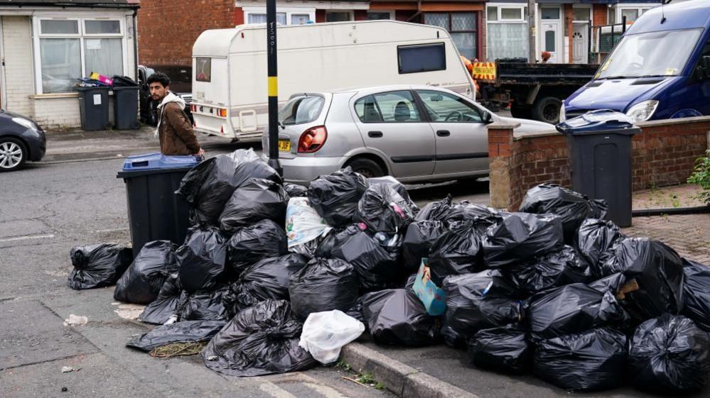 Bin bags piled up on a pavement at the junction of a residential street.