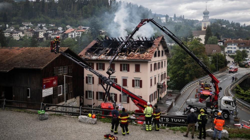 The building as seen in the daylight. Most of the roof is gone, with charred wooden beams showing. Smoke continues to rise from it. Emergency services are still at the scene - they are using large cherry picker.