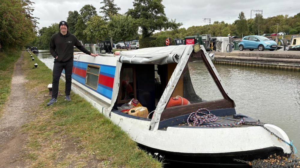 Will Retallick wearing a dark cap, dark top and dark trousers. He is standing with his left hand on a narrowboat which is moored alongside a canal towpath. The boat has a white roof and red and blue paintwork along the side. There are trees running along the towpath on both sides, and a blue car is visible in the marina on the opposite side of the canal.