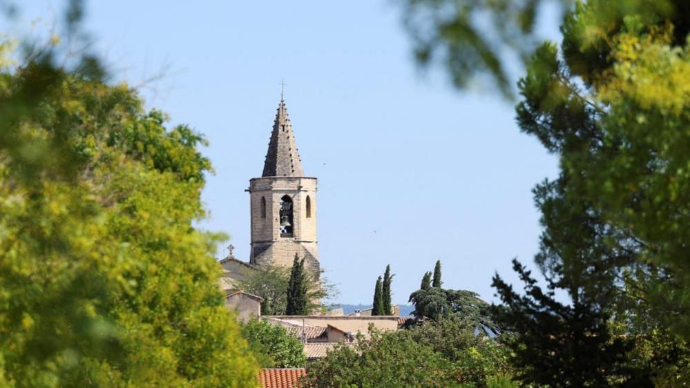 A church spire and houses seen from between green trees and undergrowth, set against a bright blue sky