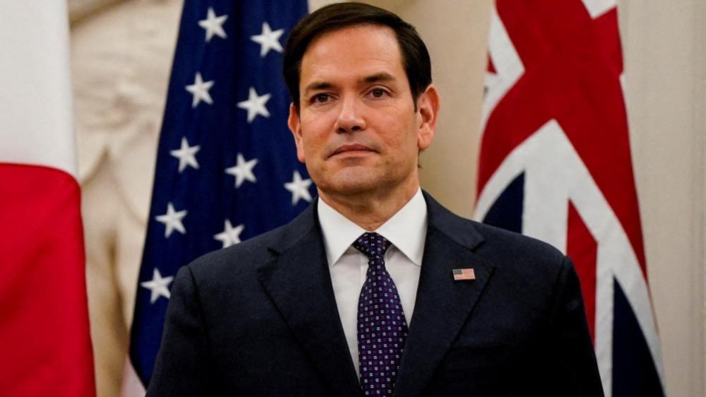 Secretary of State Marco Rubio stands in front of US and UK flags