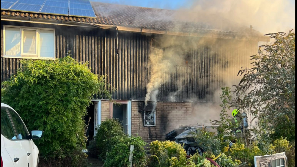 A terraced house with wooden cladding on the exterior of the first floor. Smoke is billowing from a ground floor window and the roof. A black car is in the front garden, with a firefighter standing next to it.