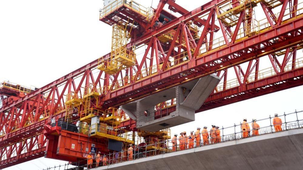 A number of workers in orange suits and hard hats stand under a red bridge.