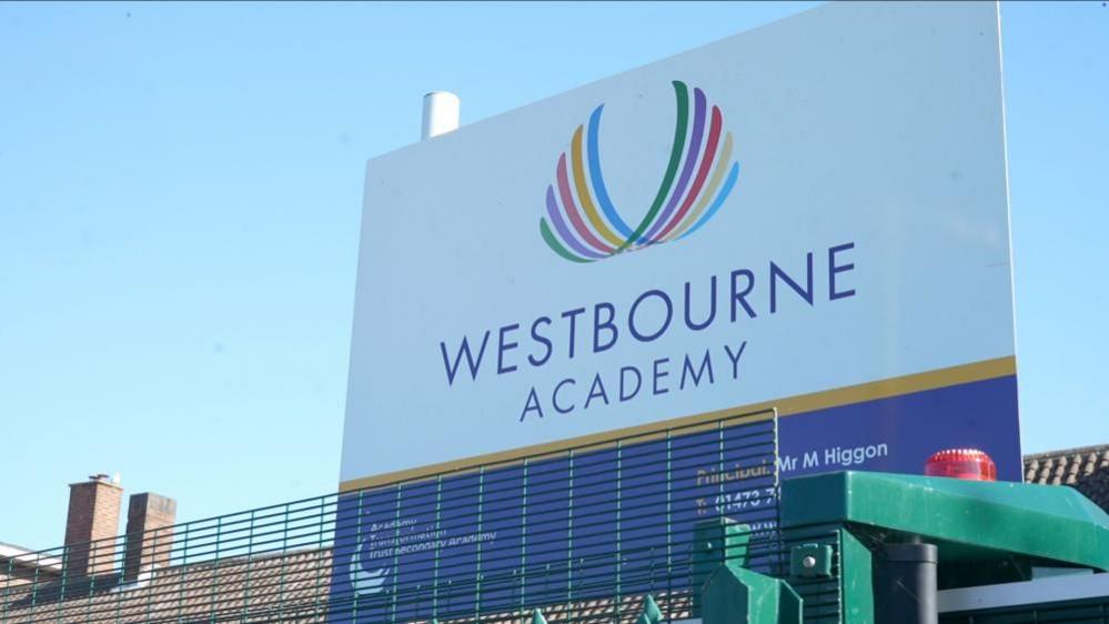 A large sign for Westbourne Academy. It is outside the school and has a colourful logo on it as well as the school name. The name of the principal, Mr Higgon and the school's telephone number are also printed on the sign