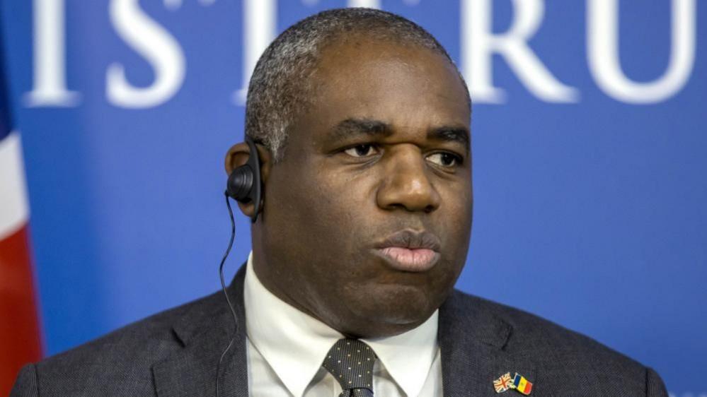 British Foreign Secretary David Lammy wearing an earpiece and a dark grey suit with a white shirt